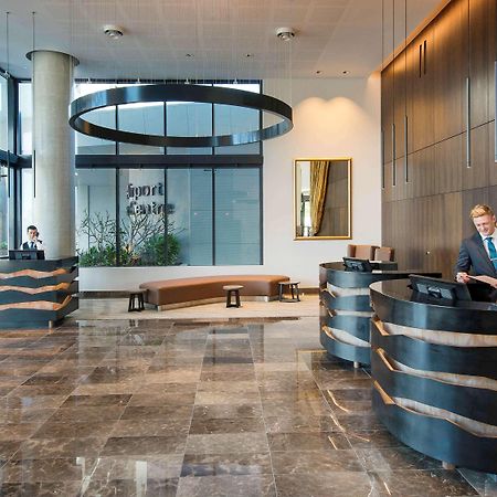 Pullman Brisbane Airport Otel Dış mekan fotoğraf The image shows a modern hotel lobby. There are two reception desks made of dark-colored materials, and staff members are attending to guests at these desks. The lobby features large windows that allow natural light to fill the space, and there are p