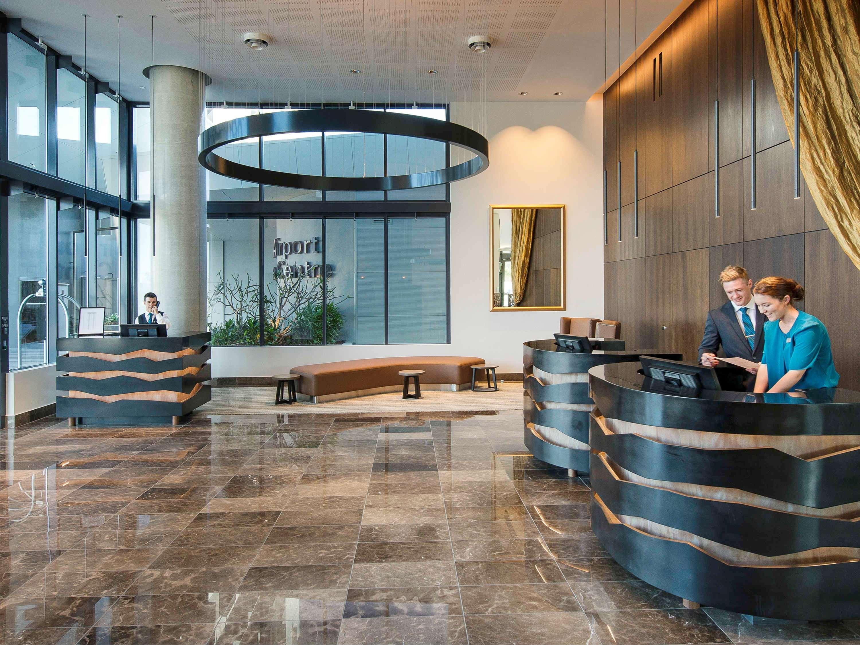 Pullman Brisbane Airport Otel Dış mekan fotoğraf The image shows a modern hotel lobby. There are two reception desks made of dark-colored materials, and staff members are attending to guests at these desks. The lobby features large windows that allow natural light to fill the space, and there are p